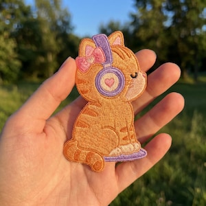 May include: An embroidered patch of an orange tabby cat wearing purple headphones with a pink heart detail and a pink bow on its head. The cat is depicted in a seated position with its eyes closed, featuring detailed stitching and a vibrant color palette.