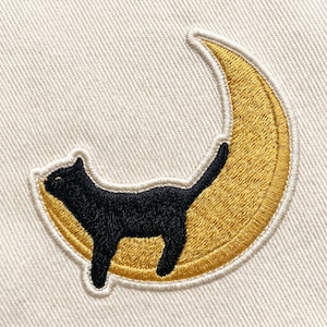 May include: Embroidered patch featuring a black cat silhouette perched on a gold crescent moon. The patch has a white border and is set against a cream-colored fabric background. The design is detailed and suitable for apparel.