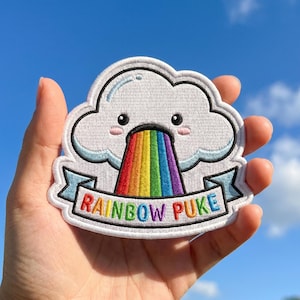May include: An embroidered patch featuring a white cloud with a smiling face and pink cheeks, vomiting a vibrant rainbow. Below the rainbow, a light blue banner displays the text RAINBOW PUKE in multicolored capital letters with a black border.