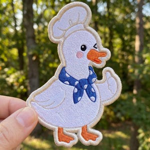 May include: Embroidered patch of a white cartoon goose wearing a chef's hat and a blue polka-dot scarf. The goose has an orange beak and feet, and is winking. The patch has a tan border and is set against a blurred green background.