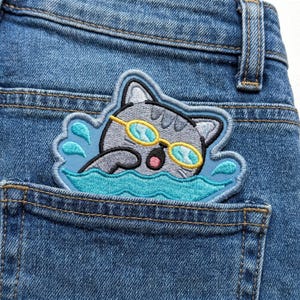 May include: Embroidered patch featuring a gray cat wearing yellow swimming goggles, swimming in blue water. The patch is sewn onto a blue denim pocket. The cat has a pink mouth and is surrounded by blue water droplets.