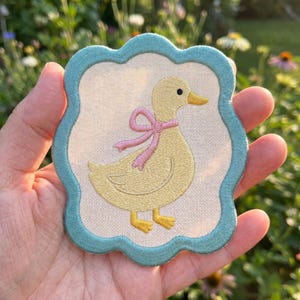 May include: Embroidered patch featuring a yellow duck with a pink bow, set against a cream background. The patch has a light blue scalloped border. The duck has orange feet and a matching beak.