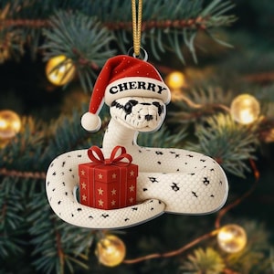 May include: A Christmas ornament featuring a white snake with black spots, wearing a red Santa hat with the name "Cherry". The snake is coiled around a red gift box with gold stars. The ornament hangs from a gold string.