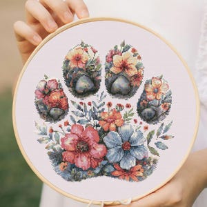 May include: A circular wooden embroidery hoop containing a cross-stitch pattern of a paw print. The paw print is composed of various colorful flowers and green leaves in shades of pink, blue, orange, and yellow against a white fabric background.