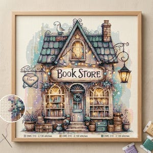 May include: A cross-stitch pattern featuring a bookstore with a teal roof, a sign reading "BOOK STORE," and a hanging lantern. The design includes shelves filled with books, a bird on the roof, and a heart-shaped sign labeled "Atore." The artwork is framed in wood and includes a circular detail showing the stitch texture.