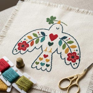 May include: A square piece of off-white fabric with a fringed edge featuring a cross-stitch design of a bird with outstretched wings, decorated with red flowers, green leaves, and small hearts. A gold thimble, embroidery floss, and scissors.