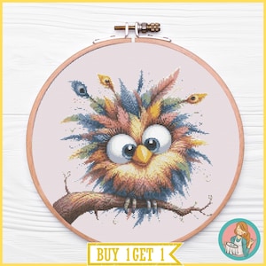 May include: A circular wooden embroidery hoop containing a cross-stitch pattern of a fluffy, wide-eyed owl with colorful feathers in shades of blue, yellow, and pink, perched on a brown branch against a light pink fabric background.