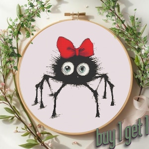 May include: Cross-stitch embroidery of a cartoon spider with a red bow. The spider is black with large blue eyes and long black legs. The embroidery is set in a wooden hoop, surrounded by greenery, with the text "buy 1 get 1".