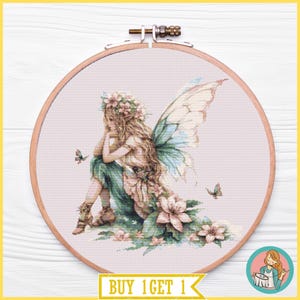 May include: A circular wooden embroidery hoop containing a cross-stitch pattern of a fairy with long hair, floral crown, and large wings, wearing a green skirt and brown footwear, surrounded by flowers and butterflies on a light pink fabric background.