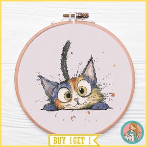 Puede incluir: Un bastidor de bordado redondo de madera que contiene un patrón de punto de cruz de un gato calicó con ojos grandes y la cola levantada. El gato está representado en tonos naranja, negro y blanco sobre un fondo de tela clara, fijado con un tornillo.
