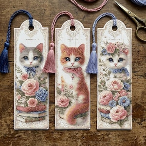 Könnte beinhalten: Drei rechteckige Kreuzstich-Lesezeichen mit Katzenmotiven. Links eine grau-weiße Katze mit Rosen und Büchern, mittig eine orangefarbene Katze mit Schal und Rosen, rechts eine weiße Katze mit Blumenkranz und Rosen, jeweils mit farbigen Quasten.