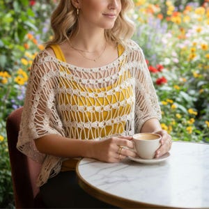 以下が含まれることがあります： クリーム色のオープンワークかぎ針編みトップスと黄色のタンクトップ。 トップスは、丸いネックラインと短い袖のゆったりとしたデザインです。 大理石のテーブルで白いコーヒーカップを持っています。