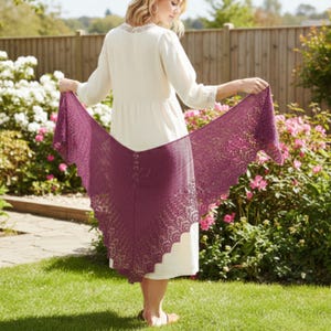 Könnte beinhalten: Eine Frau hält einen tiefvioletten Schal mit einem filigranen Lochmuster. Der Schal ist ausgebreitet und zeigt sein kompliziertes, spitzenartiges Design und die gewellten Kanten. Der Hintergrund zeigt einen Garten mit grünem Gras und rosa Blumen.