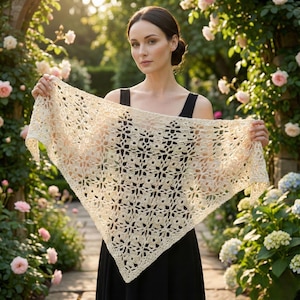 May include: A cream-colored, open-weave crochet shawl with a floral pattern. The shawl is held up, displaying its triangular shape and intricate design. The background features a garden with roses and greenery.