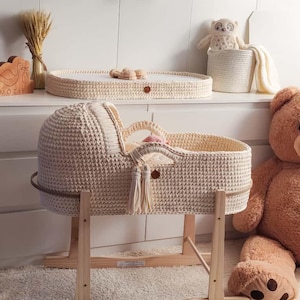 May include: A cream-colored, crocheted baby bassinet with a wooden rocking stand. The bassinet features a hood, tassels, and a handle. A matching changing table and a large teddy bear are also in the image.