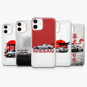 May include: A collection of phone cases featuring stylized illustrations of Nissan sports cars. Each case showcases a different car design against a backdrop of Japanese-inspired art, including a red sun, torii gates, and kanji characters. The cases are white with a red accent.