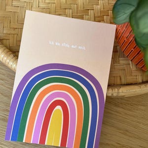 Könnte beinhalten: Eine Grußkarte mit einem Regenbogen-Design in Lila, Blau, Grün, Orange, Rosa, Rot und Gelb. Der Text "Ich bin stolz auf mich" ist in Weiß. Die Karte befindet sich auf einem hellpfirsichfarbenen Hintergrund.