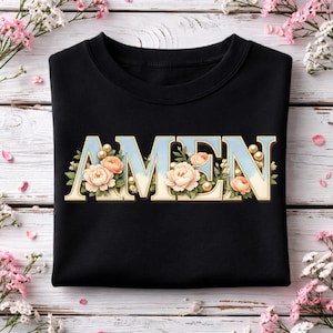 May include: Black sweatshirt featuring the word "AMEN" in a light blue gradient font, decorated with peach roses, pearls, and green leaves. The sweatshirt is folded on a white wooden surface with pink flowers.
