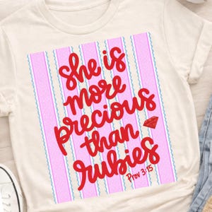 Puede incluir: Camiseta color crema con el texto rojo "She is more precious than rubies". El diseño incluye un fondo a rayas rosas y blancas, un gráfico de gema roja y el versículo bíblico Proverbios 3:15.