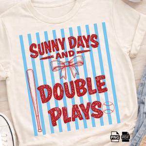 May include: A cream-coloured t-shirt with a baseball theme. The design features the text "SUNNY DAYS AND DOUBLE PLAYS" in red, along with a baseball bat, a bow, and a baseball, set against a backdrop of blue vertical stripes.