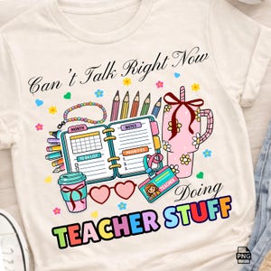 May include: Cream-colored t-shirt with a colorful graphic. The design includes a planner, pencils, a coffee cup, and the text "Can't Talk Right Now Doing TEACHER STUFF". The graphic is in bright colors with floral and heart accents.