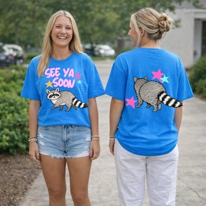 Puede incluir: Dos mujeres con camisetas azules con un diseño de mapache. La parte delantera de la camiseta dice "See Ya Soon" con estrellas. La parte trasera de la camiseta tiene un mapache y gráficos de estrellas. Una mujer lleva pantalones cortos vaqueros, la otra pantalones blancos.