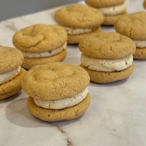 May include: Several golden-brown sandwich cookies filled with a thick, creamy white filling are arranged on a white surface. The cookies have a slightly domed shape and a soft, textured appearance, suggesting a homemade treat.