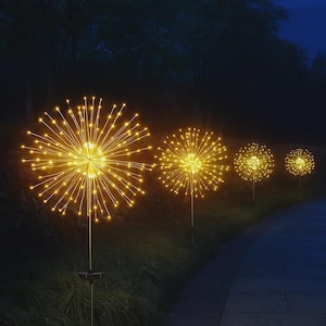 Op de afbeelding: Een nachtelijke buitenscène met een rij verlichte, bolvormige zonne-tuinverlichting. Elke lamp heeft een centrale bol met uitstralende, warmgele lichten, die op vuurwerk lijken. De lichten zijn geplaatst langs een pad en in een grasveld.