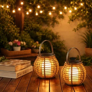 Puede incluir: Dos faroles redondos decorativos con un exterior tejido de color marrón claro y una luz brillante y cálida en el interior. Cada farol tiene un asa de metal. Los faroles están sobre una mesa de madera, con libros y plantas en el fondo.