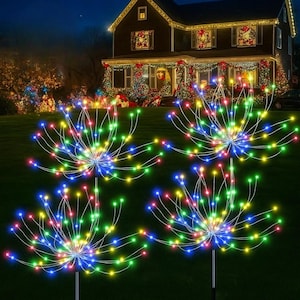 May include: Four colorful, starburst-shaped outdoor Christmas lights illuminate a dark green lawn. Each light features a central hub with radiating wires tipped with red, green, blue, and yellow LED lights. A decorated house with Christmas lights is in the background.