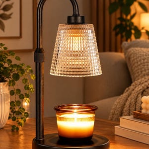 May include: A black metal candle warmer lamp with an adjustable arched arm and a textured, clear glass shade. The lamp features a circular base holding a small amber glass jar candle that is illuminated with a warm, glowing light from above.