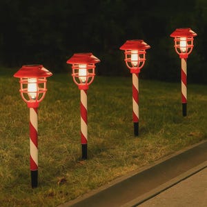 Puede incluir: Cuatro luces solares para jardín. Cada luz tiene una parte superior y una jaula rojas, un poste a rayas blancas y rojas y una base negra. Las luces están dispuestas en fila sobre un césped, con un fondo oscuro.