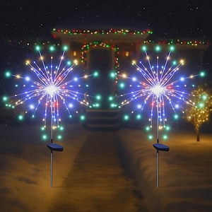 Può includere: Due luci solari da esterno a forma di stella, con luci a LED blu, verdi e rosse. Le luci sono su pali di metallo con pannelli solari e sono posizionate in un cortile innevato. Sullo sfondo c'è una casa con luci natalizie.