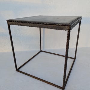 Può includere: Un tavolino quadrato con un piano grigio scuro e una struttura in metallo arrugginito. Il piano del tavolo ha un bordo strutturato e le gambe sono realizzate con tondini di metallo ritorti. Questo tavolo in stile industriale è adatto per uso interno o esterno.