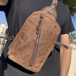 Könnte beinhalten: Eine braune Canvas-Umhängetasche mit vertikalem Reißverschluss und Lederakzenten. Die Tasche wird über der Brust getragen und hat einen Schultergurt. Die Tasche hat ein robustes, abgenutztes Aussehen.