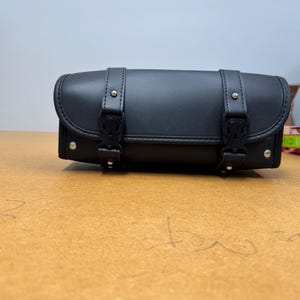 May include: A black leather motorcycle bag with two adjustable straps and buckles. The bag is rectangular with rounded edges and silver rivets. It is sitting on a light brown surface.