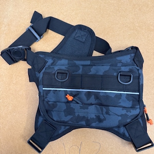 May include: Black and blue camouflage fanny pack with adjustable straps and multiple compartments. Features a reflective stripe and orange accent cords. Includes a black buckle closure and D-rings for attachments.