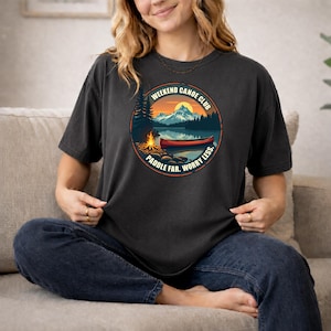 Puede incluir: Una camiseta de manga corta gris carbón con un gráfico circular de un paisaje montañoso al atardecer con una fogata y una canoa roja en un lago. El texto dice WEEKEND CANOE CLUB y PADDLE FAR. WORRY LESS. en un borde circular.