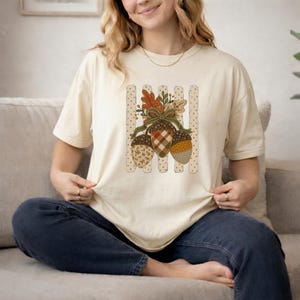 Puede incluir: Una camiseta de manga corta color crema con un gráfico central de tres bellotas con patrones variados que incluyen cuadros, flores y lunares. Las bellotas están atadas con una cinta verde y adornadas con hojas de otoño.