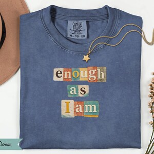 Peut inclure: Un t-shirt bleu denim avec les mots "enough as I am" dans une police colorée de style collage. Un collier pendentif étoile dorée et un chapeau marron sont visibles. Le t-shirt a une étiquette Comfort Colors.
