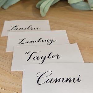 Puede incluir: Cuatro tarjetas de mesa blancas con elegante caligrafía negra. Los nombres "Sandra", "Lindsay", "Taylor" y "Cammi" están escritos en las tarjetas. Las tarjetas están dispuestas sobre una superficie de madera clara, con vegetación en el fondo.