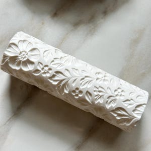 May include: A cylindrical white clay rolling pin featuring a deeply embossed pattern of intricate flowers and leafy vines across its entire surface. The tool has a smooth, matte finish and is designed for creating detailed impressions in soft materials.