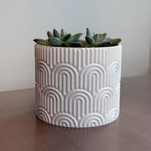 May include: A cylindrical white planter with a textured, repeating arch design, holding two green succulents. The planter sits on a brown surface, creating a contrast with the light-colored pot and plants.