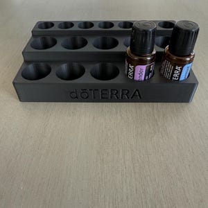 May include: A black essential oil organizer with multiple circular slots for bottles. The organizer has two levels and the brand name "dōTERRA" is printed on the front. Two essential oil bottles are in the organizer.