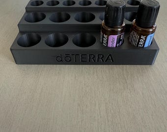 May include: A black essential oil organizer with multiple circular slots for bottles. The organizer has two levels and the brand name "dōTERRA" is printed on the front. Two essential oil bottles are in the organizer.