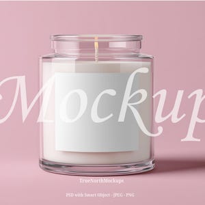 May include: A clear glass jar candle with a white label, filled with white wax. The candle has a wick and is set against a pink background. The word "Mockup" is written across the image in a cursive font.