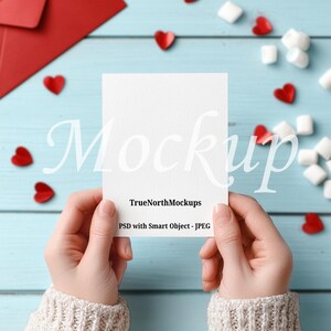 May include: A white card held by hands, with the word "Mockup" in a script font. The card is surrounded by red heart-shaped confetti, marshmallows, and a red envelope on a light blue wooden surface. The text "TrueNorthMockups" and "PSD with Smart Object - JPEG" are also visible.