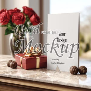 Könnte beinhalten: Ein Mockup einer Karte oder Einladung mit dem Text "Your Design Here". Eine rote Geschenkbox mit goldener Schleife steht neben der Karte, zusammen mit zwei Schokoladentrüffeln. Im Hintergrund eine Vase mit roten Rosen.