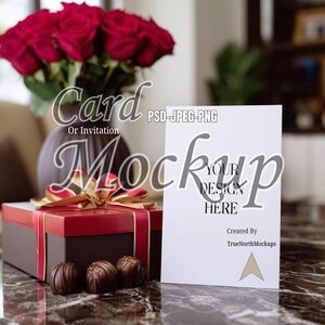 Könnte beinhalten: Ein Mockup mit einer weißen Karte mit dem Text „YOUR DESIGN HERE“ und einer rot-braunen Geschenkbox mit goldenem Band. Eine Vase mit roten Rosen und Schokoladentrüffeln sind ebenfalls vorhanden. Die Karte ist für eine Karte oder Einladung.