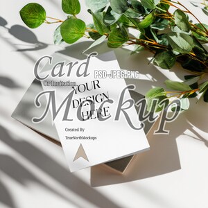 Peut inclure: Maquette de carte ou d'invitation avec le texte « VOTRE DESIGN ICI » et « Carte ou Invitation » dans une police stylisée. La carte est posée sur une enveloppe grise et une petite boîte marron clair. Des feuilles vertes sont en arrière-plan.